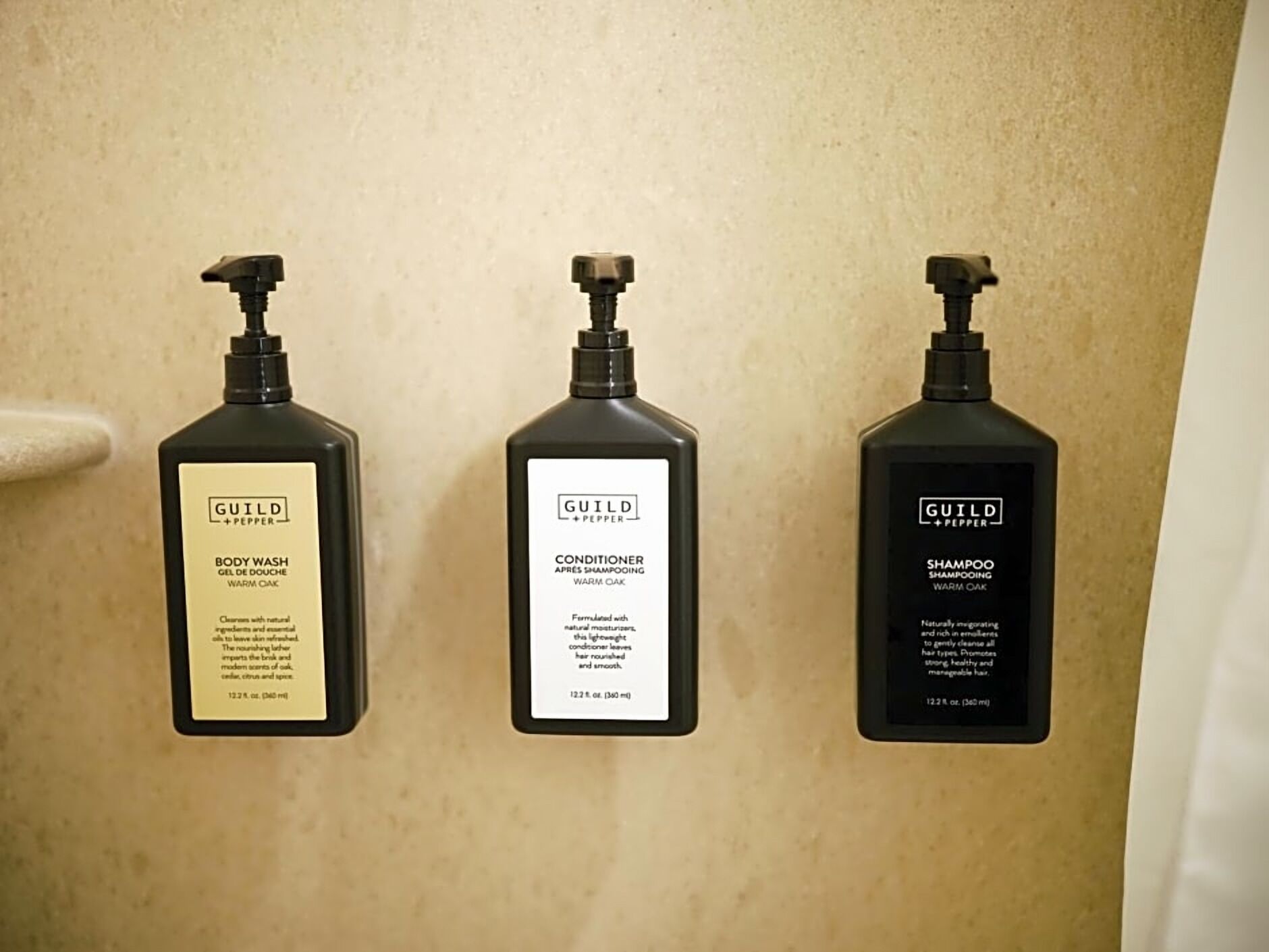 Tres botellas de crema limpiadora, acondicionador y champú Guild + Pepper sobre una pared color beige.