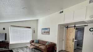 A spacious living room with a brown leather sofa, a coffee table, and a TV. The room has wooden flooring and a sliding barn door leading to a bathroom.