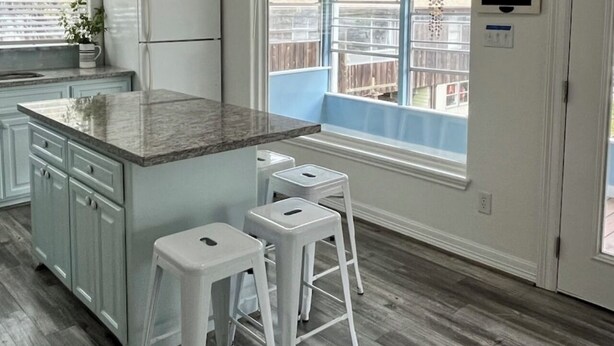 A kitchen with a granite countertop, white cabinets, a refrigerator, and a window with a view of a wooden deck.