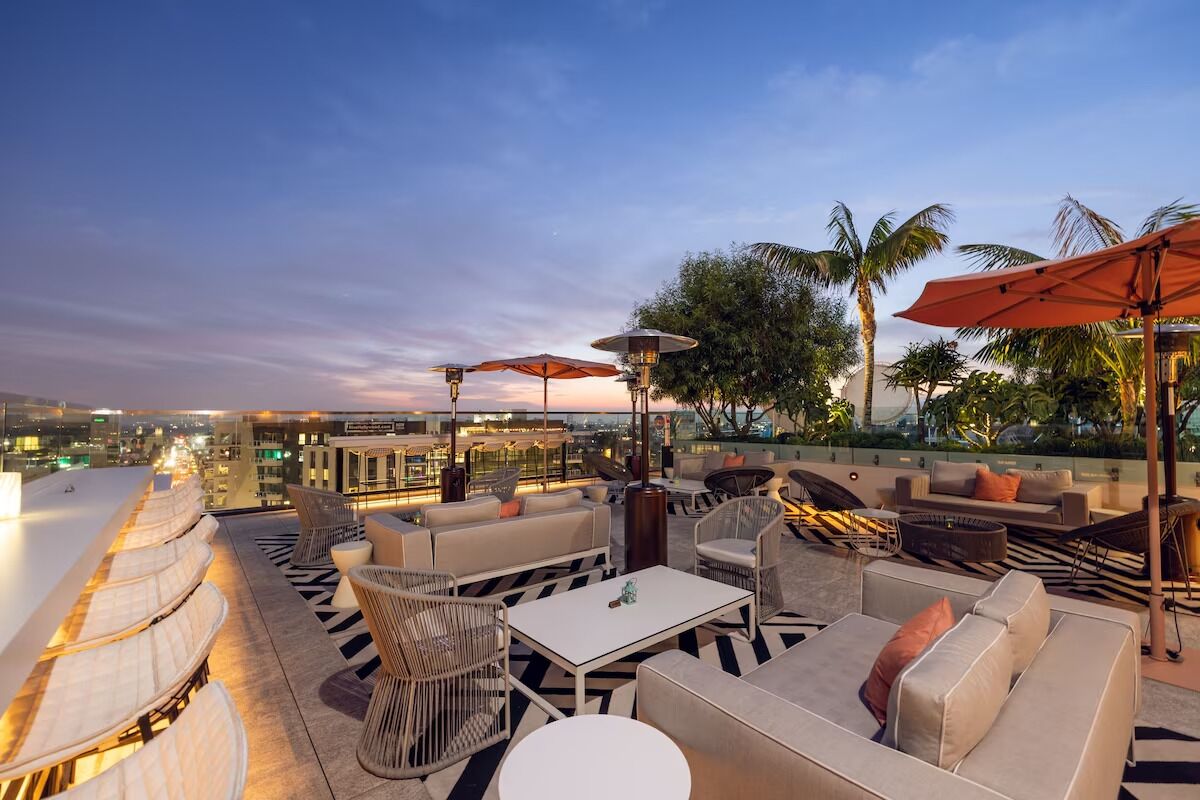 A rooftop lounge with outdoor seating, umbrellas, and a city view at dusk.