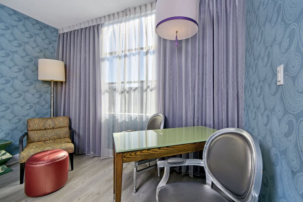 Een kamer met een blauwe wand met patroon, een houten eettafel, een rode poef, een fauteuil met patroon en een lamp.