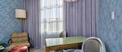 Een kamer met een blauwe wand met patroon, een houten eettafel, een rode poef, een fauteuil met patroon en een lamp.
