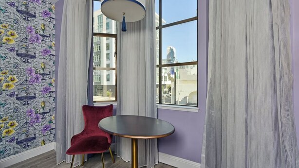 Une pièce avec un mur violet, un papier peint à motifs, une chaise rouge, une table ronde et une vue sur les bâtiments à travers de grandes fenêtres.