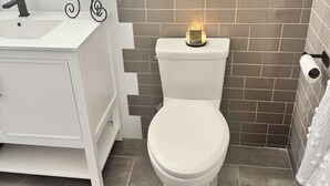 Un baño con un lavamanos blanco, un espejo, un inodoro y una vela encendida sobre el inodoro.