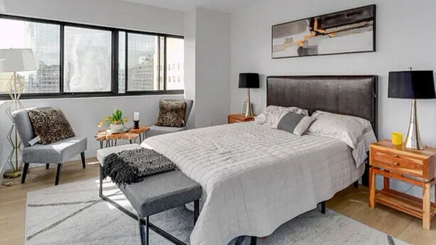 Un dormitorio moderno con una cama grande, mesitas de noche, un banco y vistas de la ciudad a través de amplias ventanas.