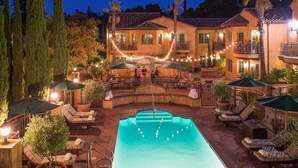 A pool area with lounge chairs and umbrellas, surrounded by a building with multiple balconies and string lights.