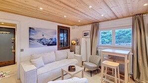 A cozy living room with a sofa, armchair, and a small table. There's a painting of mountains on the wall, a window with a view of trees, and a door with a handle.