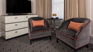 Une chambre d’hôtel avec deux fauteuils gris agrémentés de touches de couleur orange, un téléviseur à écran plat posé sur un meuble blanc et une moquette à motifs.