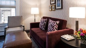 Habitación de hotel con sofá de cuero marrón, un sillón a juego, una mesita con un atril de artículos de aseo y una ventana con persianas.