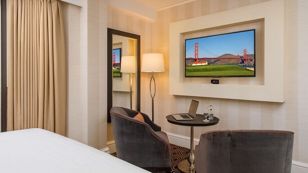 Ett hotellrum med en säng, två fåtöljer, ett skrivbord med en bärbar dator och ett glas vatten, en lampa, en tv som visar Golden Gate-bron och en spegel.
