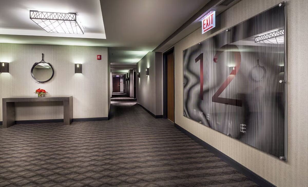 A hotel hallway with a patterned carpet, a reception desk with a vase of flowers, wall sconces, and an exit sign.