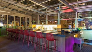 A bar area with a long counter and red chairs, large windows, and a city view.
