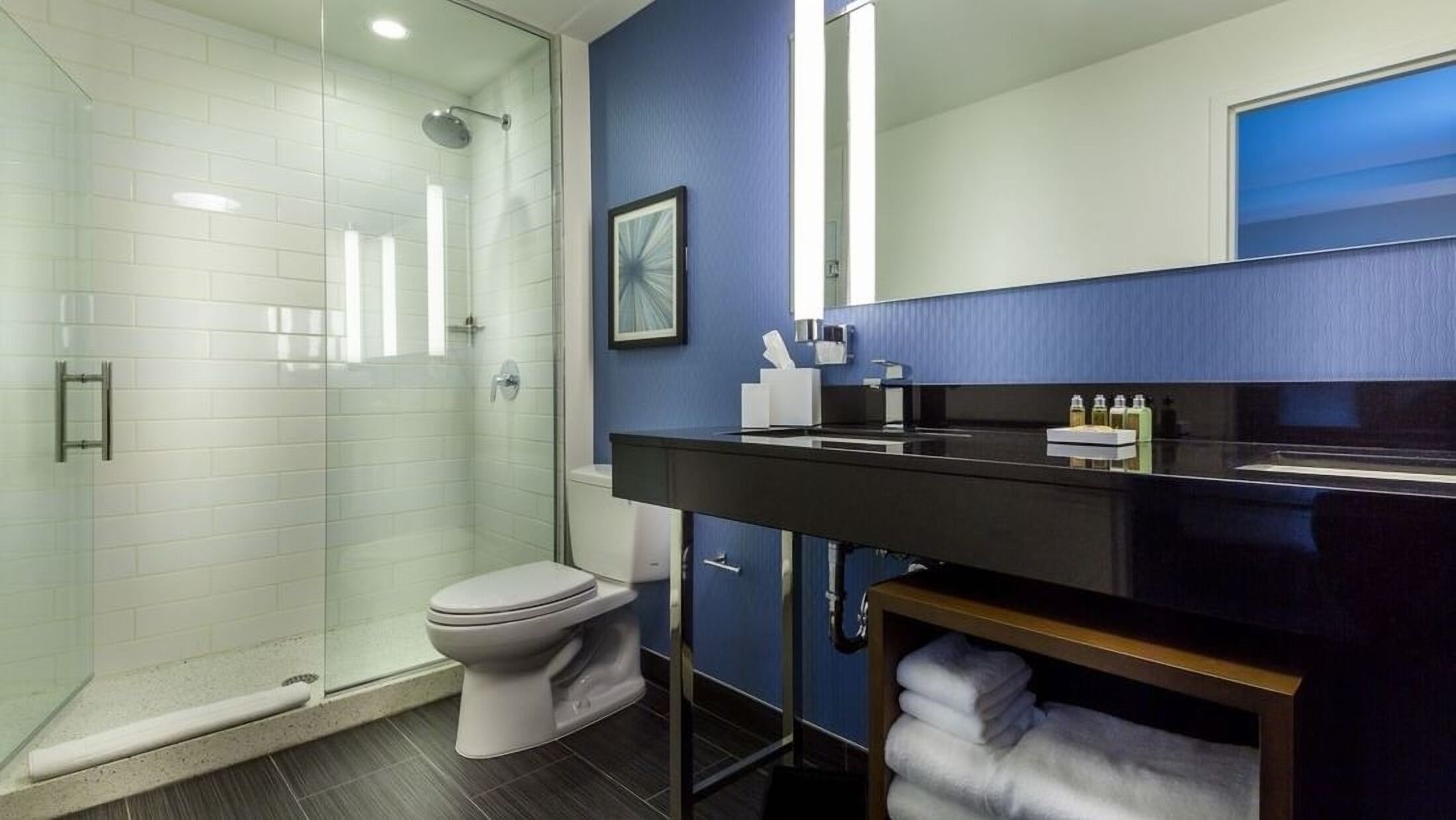 A modern bathroom with a glass-enclosed shower, a toilet, a black vanity with a mirror, and a blue accent wall.