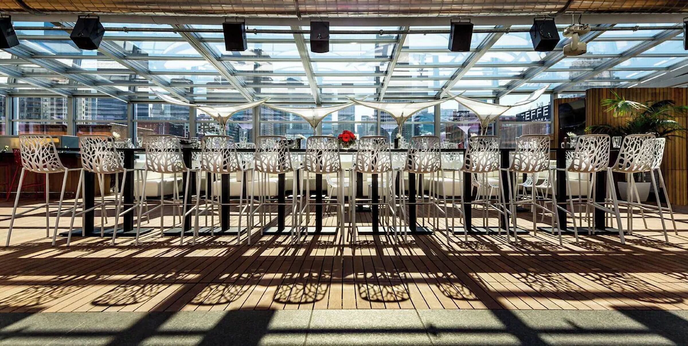 Un área de comedor al aire libre con sillas y mesas blancas, techo de vidrio y vista panorámica de la ciudad.