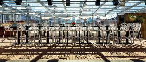 Un área de comedor al aire libre con sillas y mesas blancas, techo de vidrio y vista panorámica de la ciudad.