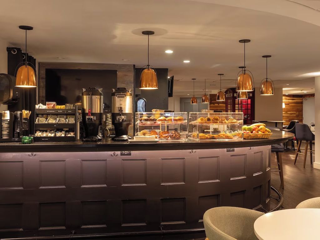 Eine moderne Bar mit einer Theke, auf der verschiedene Speisen präsentiert werden, Pendelleuchten und einer Sitzgelegenheit mit runden Tischen.