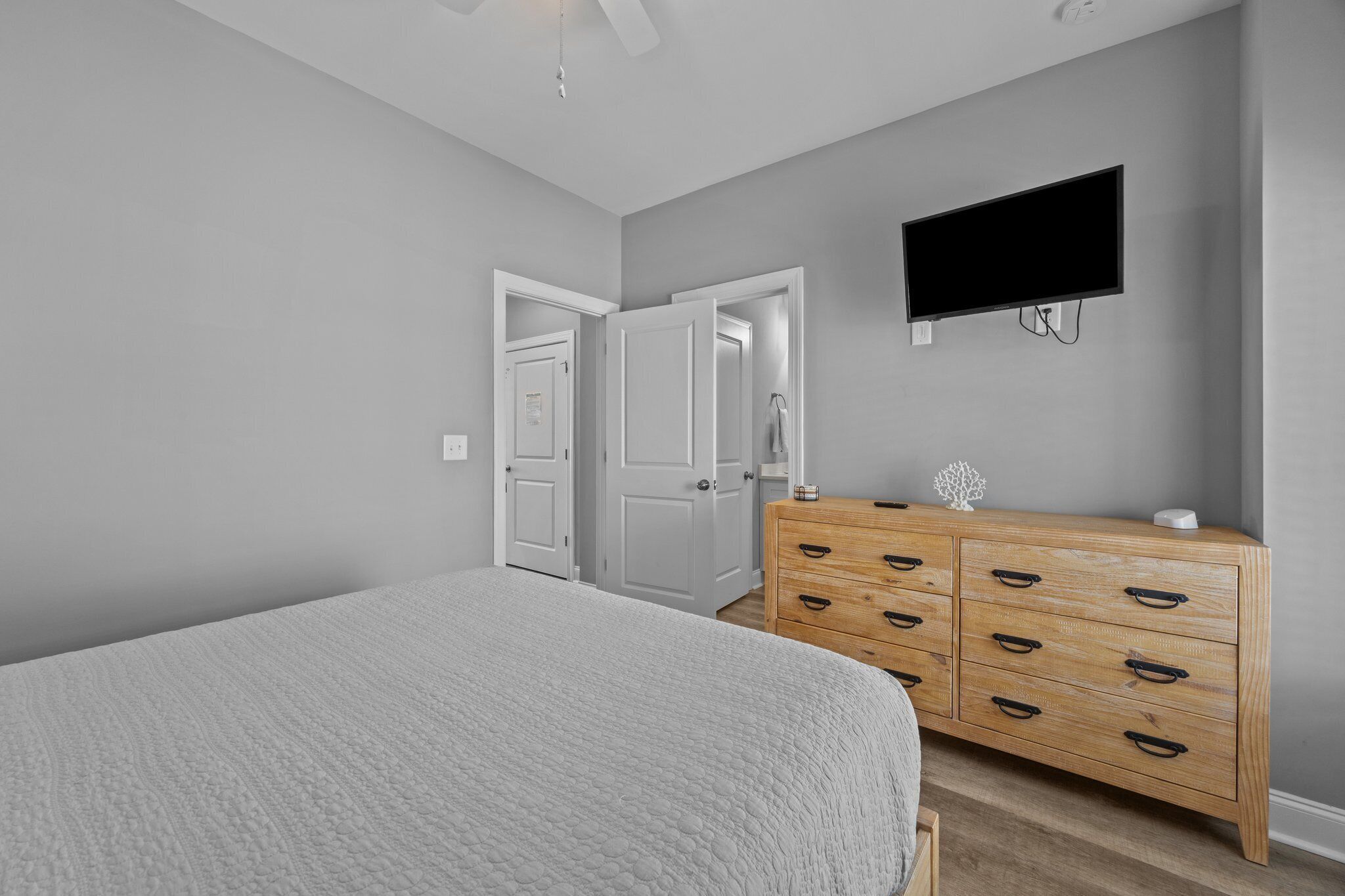 A bedroom with a bed, a wooden dresser, a television mounted on the wall, and a door leading to another room.