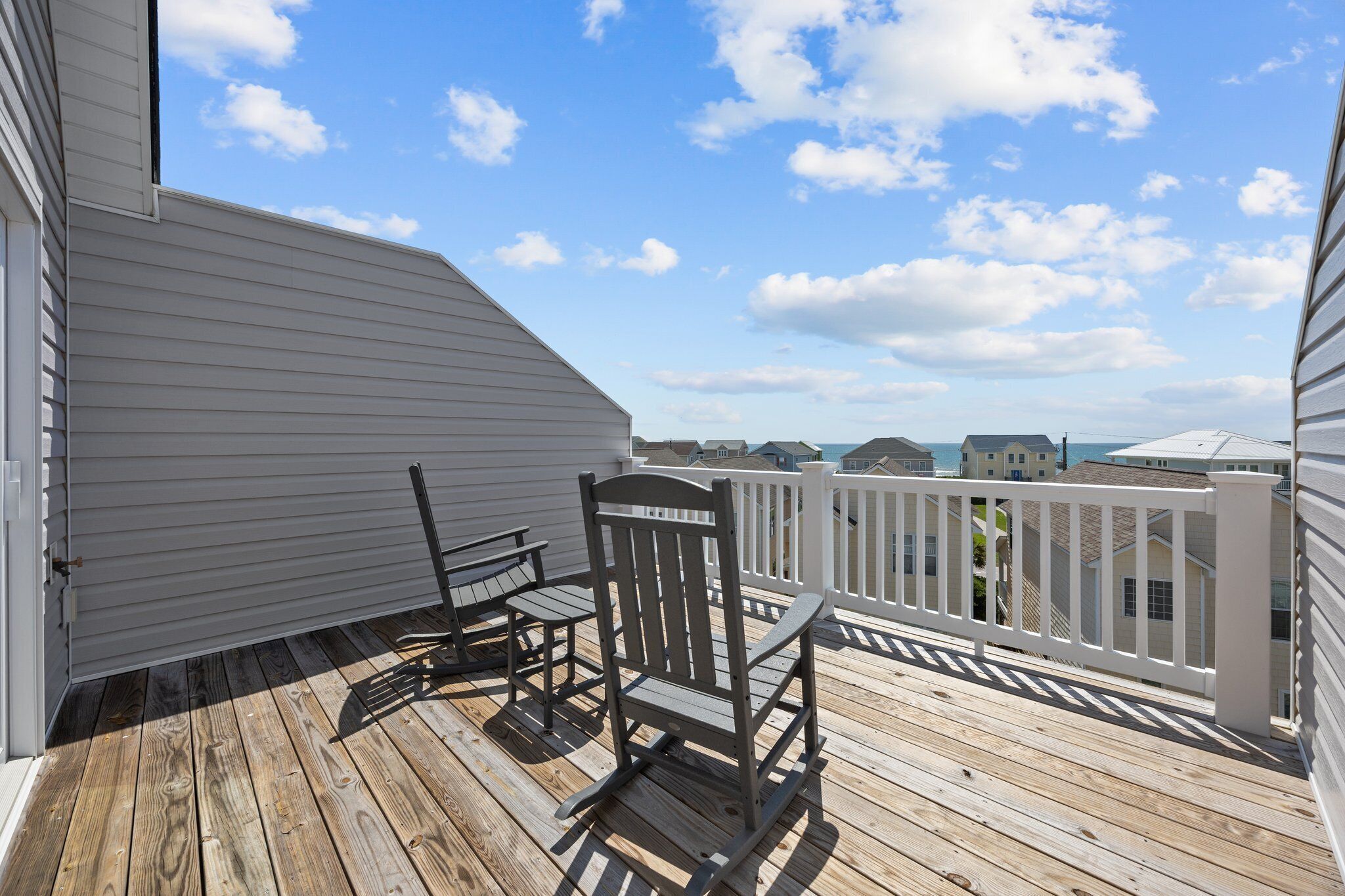 A wooden deck with a rocking chair and a small table, overlooking a residential area with houses and a clear sky.