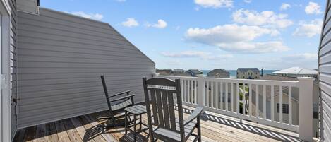 A wooden deck with a rocking chair and a small table, overlooking a residential area with houses and a clear sky.