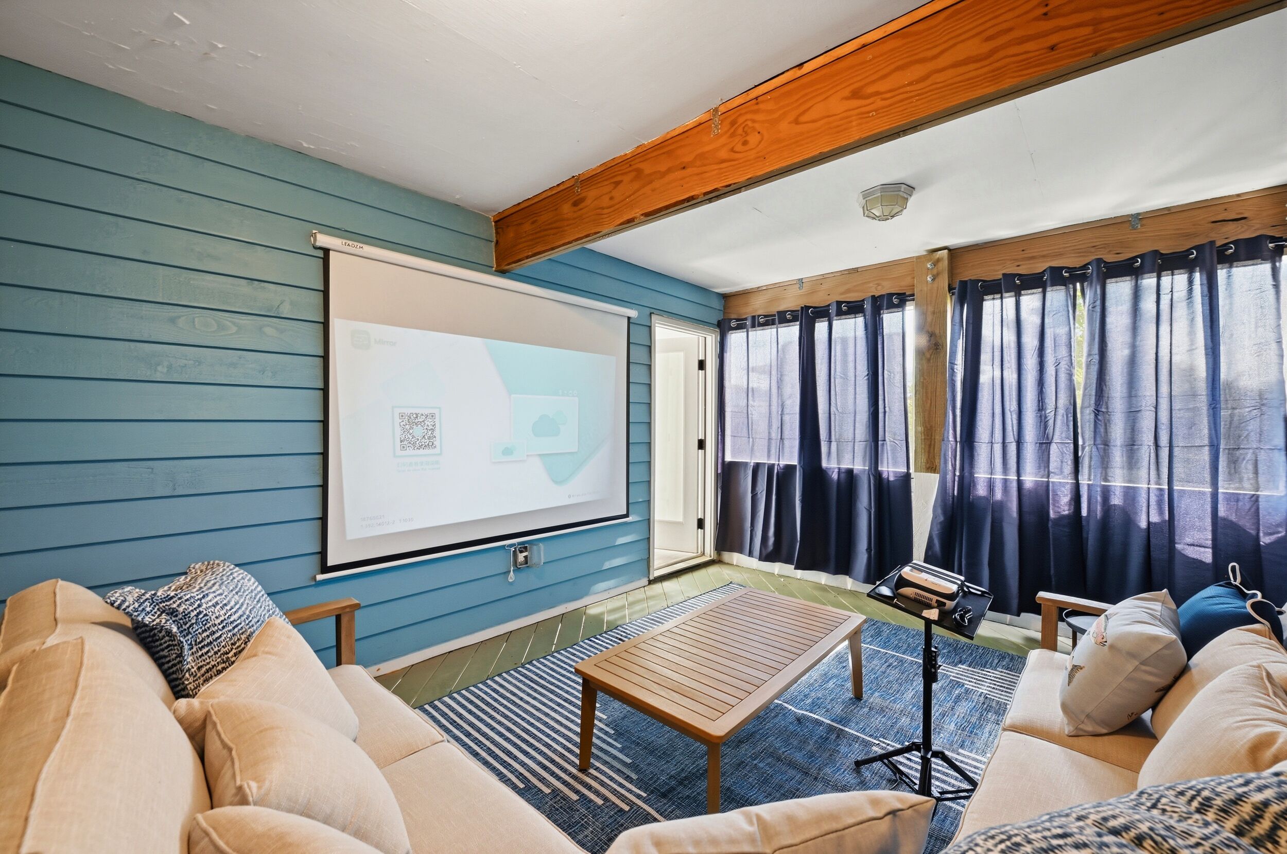 A room with a blue wall, a wooden ceiling beam, a projector screen, a sofa, a coffee table, and a window with curtains.