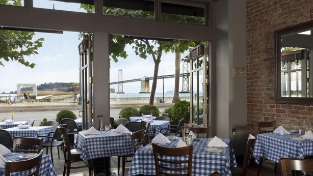 An outdoor dining area with checkered tablecloths and wooden chairs, overlooking a river with a bridge in the background. - European Room Near Ghirardelli Square, Civic Center & Ferry Terminal (San Francisco)
