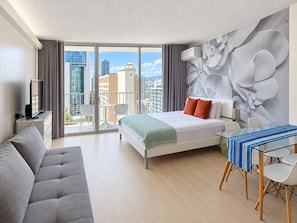 A modern hotel room with a balcony, a bed with a patterned bedspread, a glass dining table, and a grey sofa.