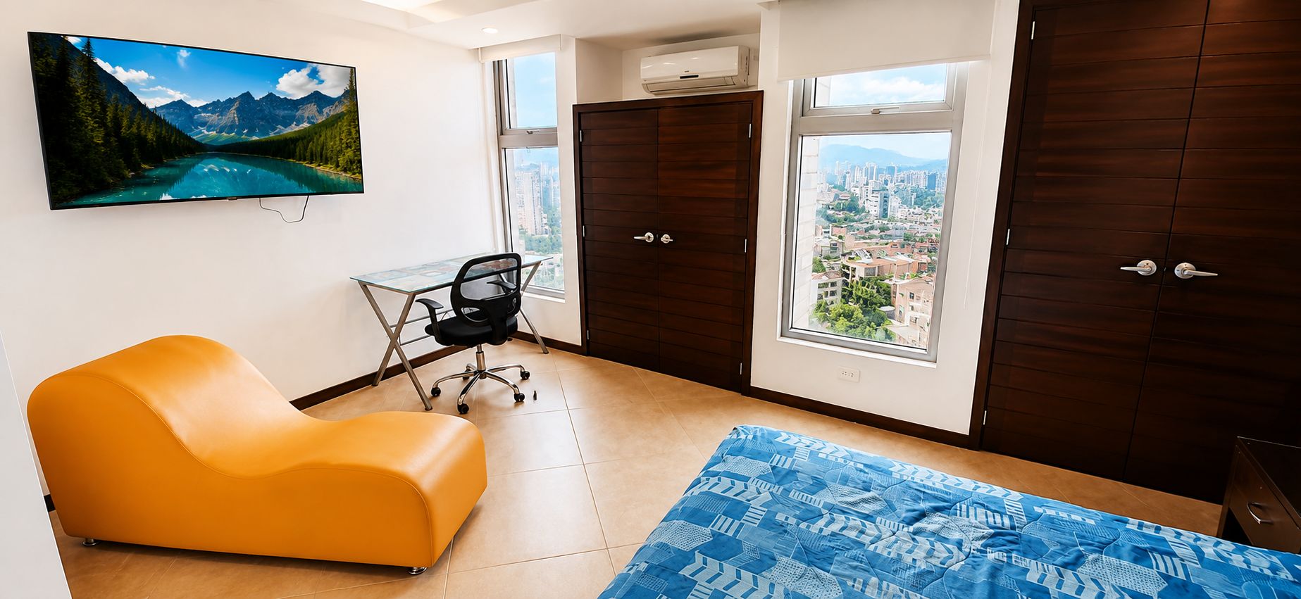 Una habitación con un ventanal grande, un escritorio, una silla naranja y una cama con colcha azul estampada.