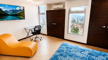 Una habitación con un ventanal grande, un escritorio, una silla naranja y una cama con colcha azul estampada.