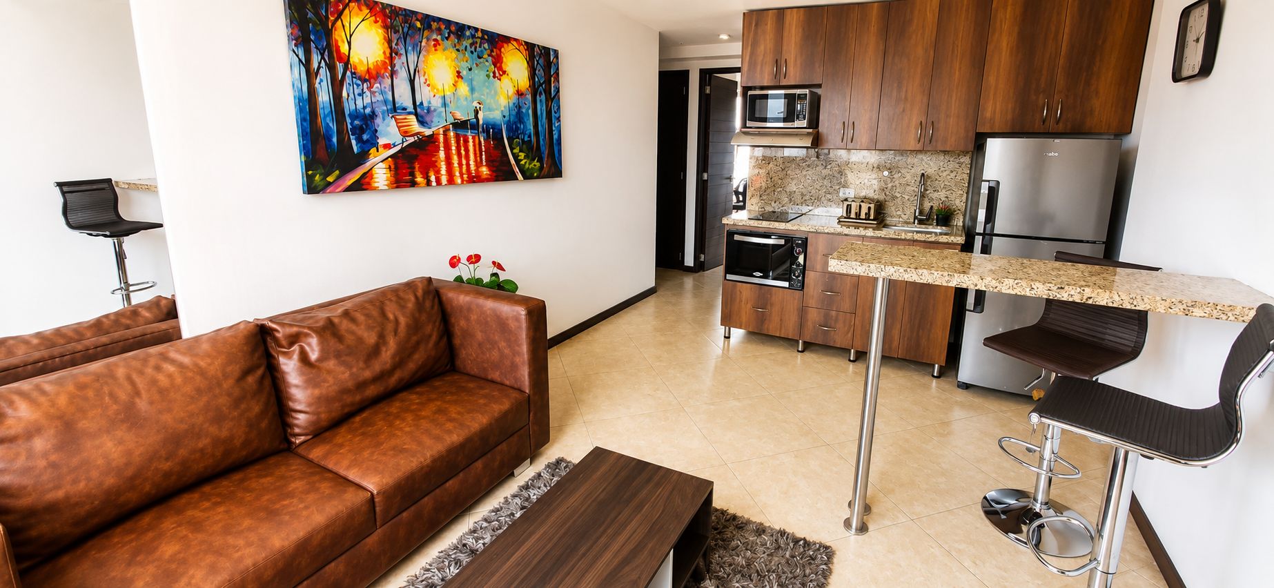 Cocina moderna con microondas, horno y refrigerador. En la sala de estar, hay un sofá de cuero marrón y un taburete de bar.