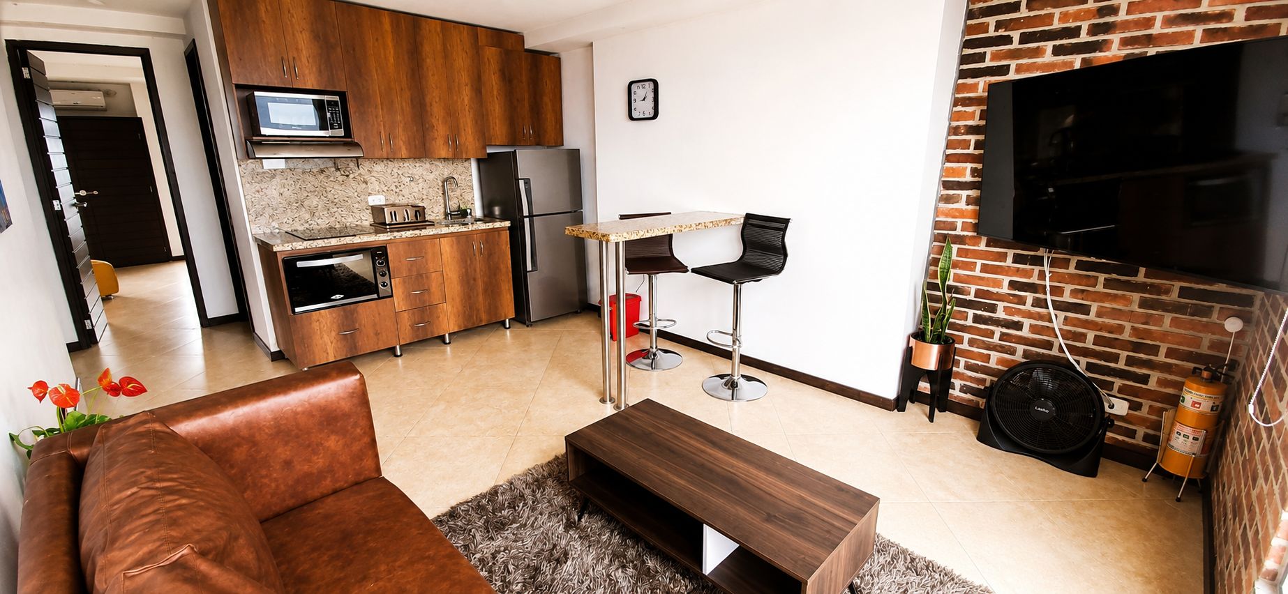 Una cocina moderna con armarios de madera, un refrigerador de acero inoxidable y un microondas. Hay una zona de comedor con una mesa y dos sillas. Un televisor de pantalla plana está montado en una pared de ladrillo.