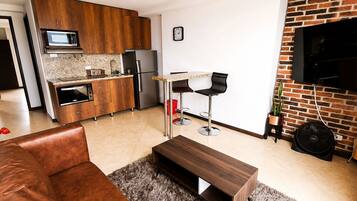 Una cocina moderna con armarios de madera, un refrigerador de acero inoxidable y un microondas. Hay una zona de comedor con una mesa y dos sillas. Un televisor de pantalla plana está montado en una pared de ladrillo.