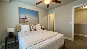 A bedroom with a bed, a nightstand, a lamp, a ceiling fan, a painting of a lighthouse, and a door leading to another room.