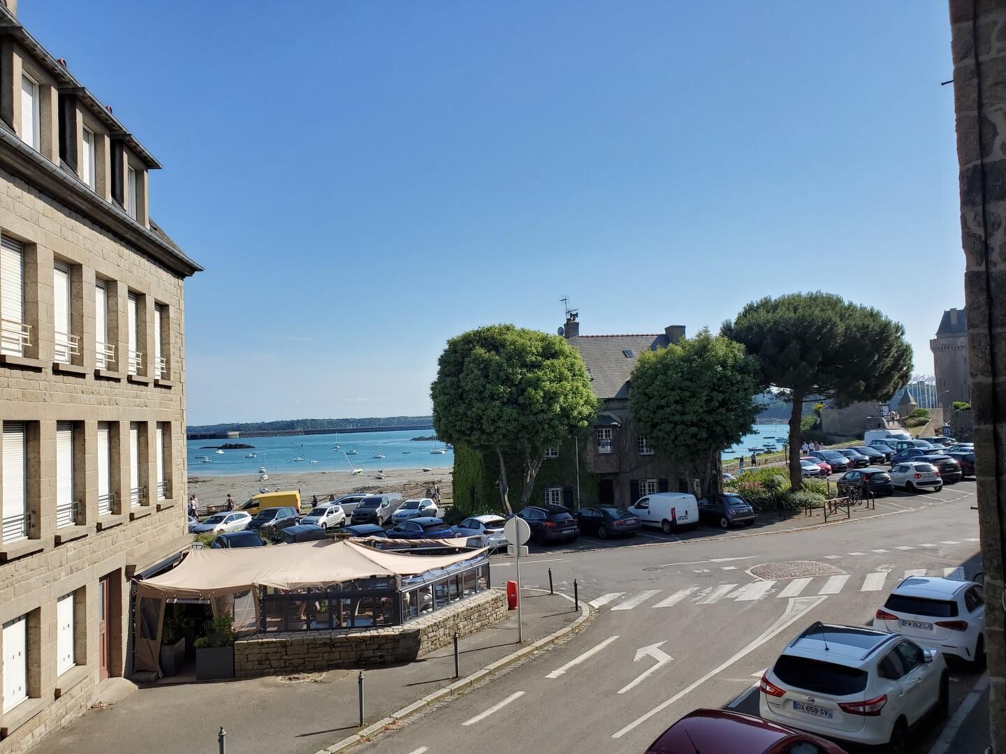 Eine Küstenstraße mit parkenden Autos, ein Café mit Außenbestuhlung und Blick aufs Meer.