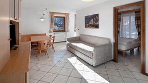 A room with a sofa, a dining table with chairs, a window with curtains, a painting on the wall, and a bed with a canopy.