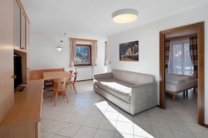 A room with a sofa, a dining table with chairs, a window with curtains, a painting on the wall, and a bed with a canopy.