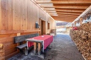 Un pasillo de madera con un banco y una mesa, una pared de piedra con leña apilada, y un edificio de madera al fondo.