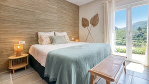A bedroom with a bed, bedside lamps, a bench, a view of greenery, and a ceiling fan.