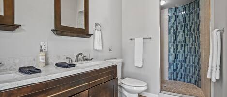 Un baño con lavamanos de mármol, tocador de madera oscura, inodoro blanco y ducha con mosaicos azules.