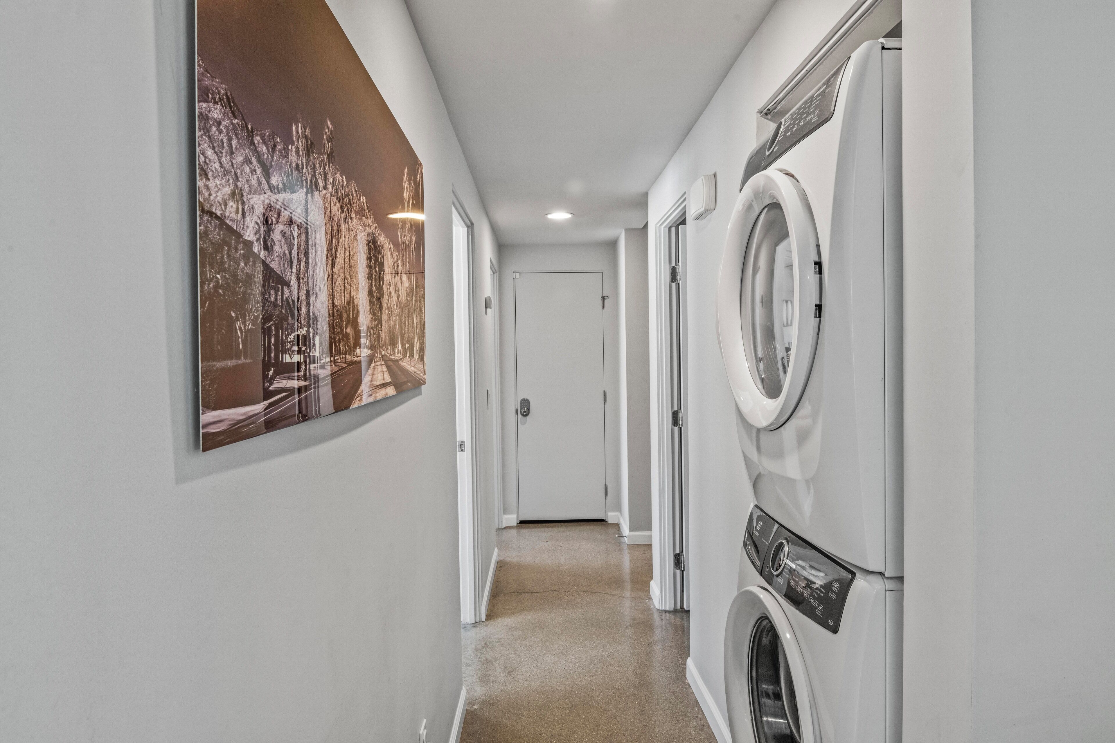 Un cuarto de lavandería con lavadora y secadora, una puerta al final del pasillo y un cuadro grande en la pared.