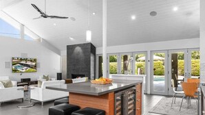 A modern kitchen with a central island, bar stools, a ceiling fan, and a large window overlooking a garden.
