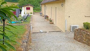 Un sentiero sterrato conduce a una casa con tetto in tegole e pareti beige. Sono presenti sedie da esterno e ombrelloni, e un piccolo giardino con vasi di piante.