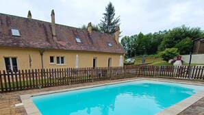 Una piscina rettangolare con bordo piastrellato, circondata da una recinzione in legno e una casa con tetto e camini in tegole.