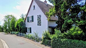 A two-story house with a white facade and a tiled roof, surrounded by greenery and a metal fence along the street. - Wellness oasis in a wonderful panoramic location (Weinheim)