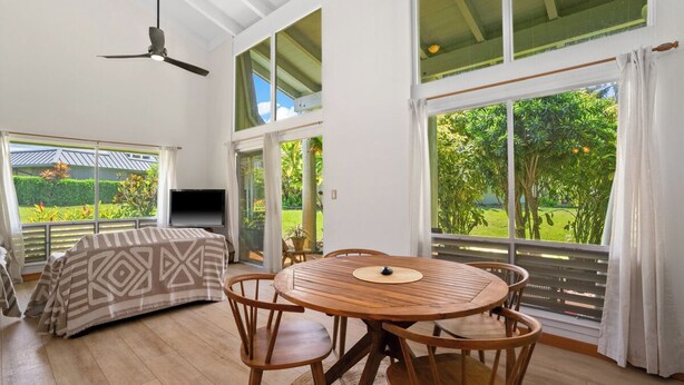 A spacious room with a wooden dining table, chairs, and a ceiling fan. Large windows offer views of a garden. - Charming 2-bedroom condo with AC, WiFi in delightful Princeville (Princeville)
