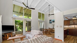 A spacious living area with a sofa, a coffee table, and a TV. There is a dining area with a table and chairs. The room has large windows and a ceiling fan. - Charming 2-bedroom condo with AC, WiFi in delightful Princeville (Princeville)