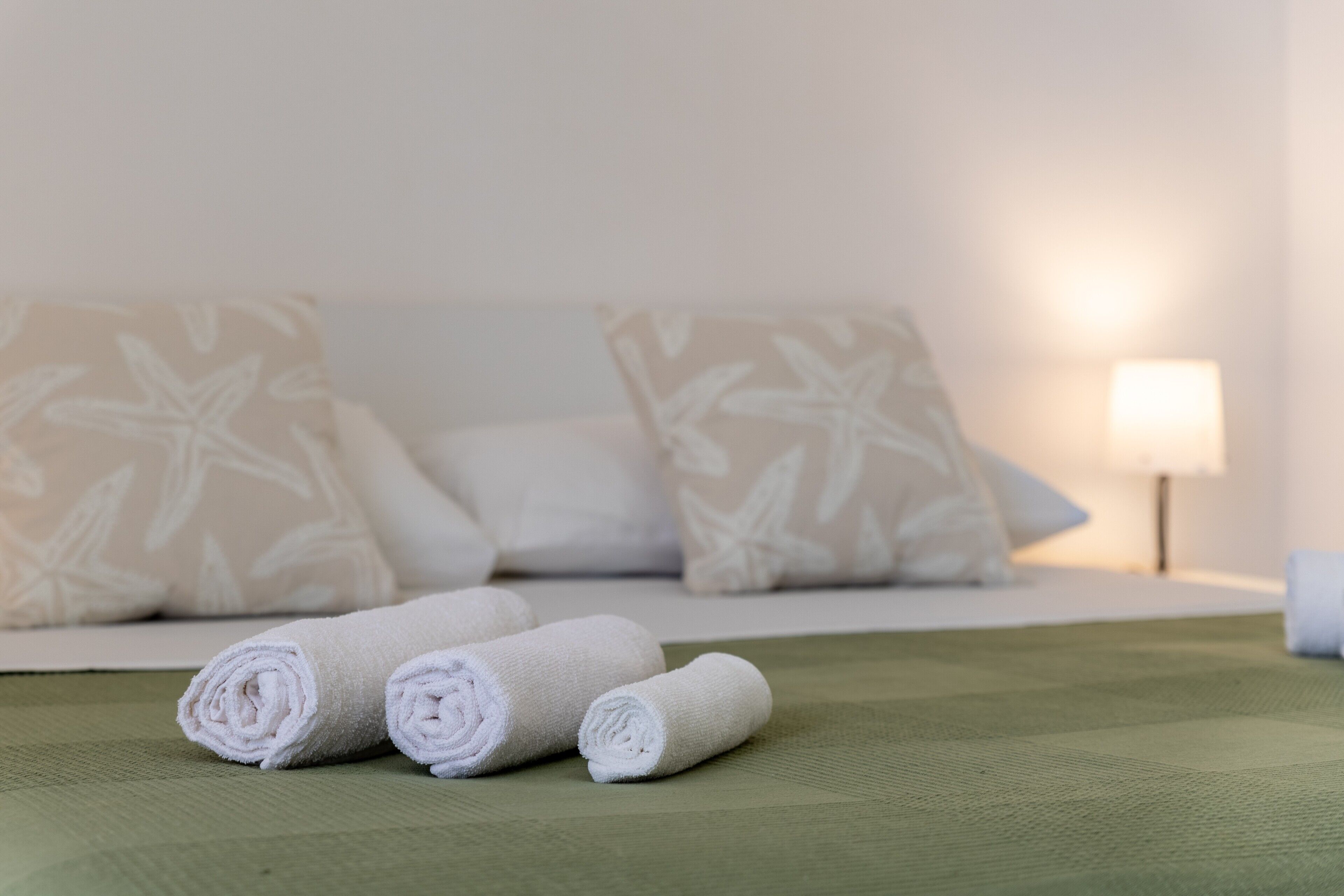Ein ordentlich bezogenes Bett mit aufgerollten weißen Handtüchern, einem grünen Bettüberwurf und einer Lampe auf einem Nachttisch.