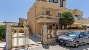Un edificio de dos pisos con entrada protegida por una reja, un coche estacionado adelante y una palmera.