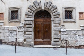 A stone building with a large wooden double door, two small windows with metal grilles, and a stone wall with carvings.