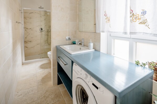 A bathroom with a glass shower enclosure, a sink with a faucet, a washing machine, and a mirror.