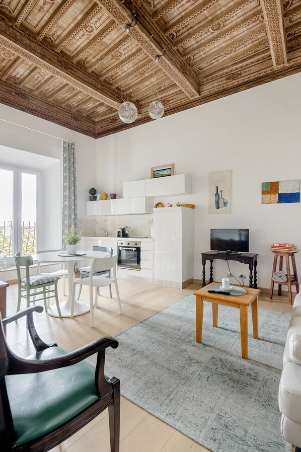 A modern living room with a wooden ceiling, a dining area with chairs, a kitchen with white cabinets, a television, and artwork on the wall.